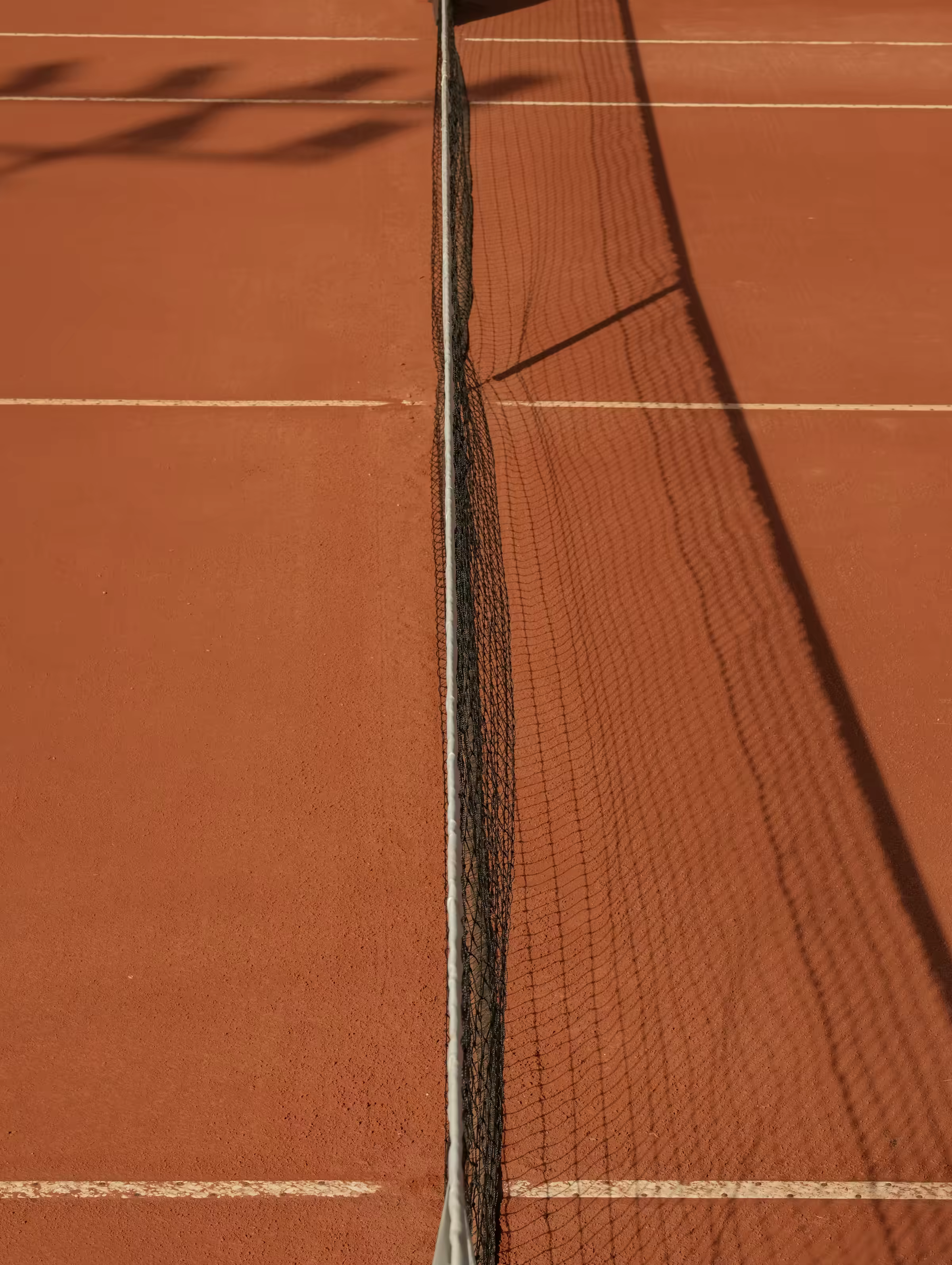 A tennis net casting a long shadow across a red clay court at golden hour.